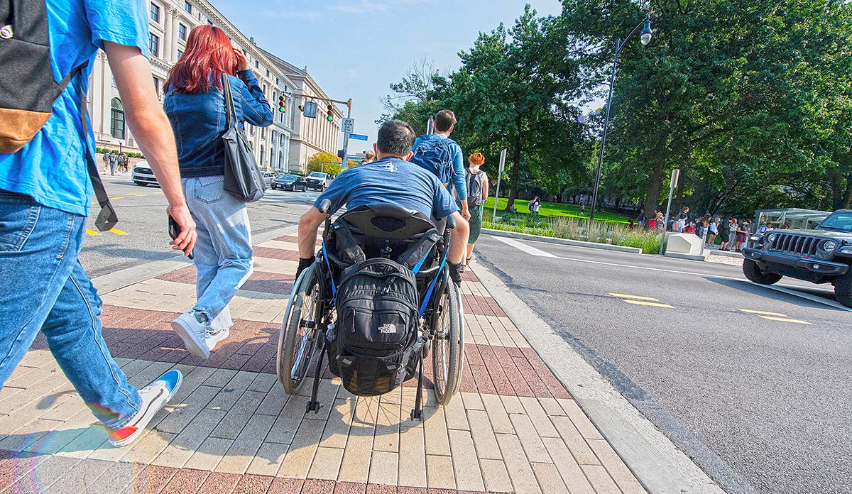 A person using a wheelchair crosses Bigelow Boulevard between the Cathedral of Learning and William Pitt Union