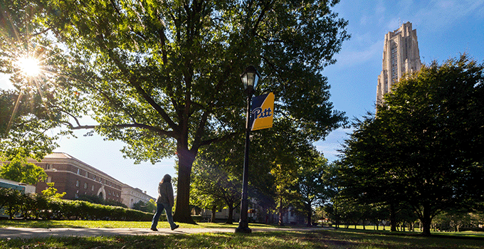 Person walking on campus with trees and the Cathedral of Learning in the background