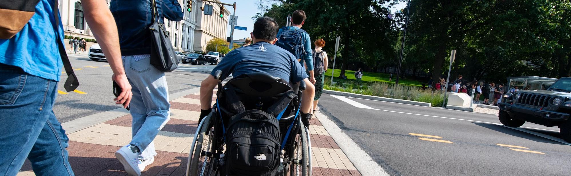 A picture of a group of people crossing Bigelow Boulevard on the Pittsburgh campus. The central focus is on a person crossing in their wheelchair.
